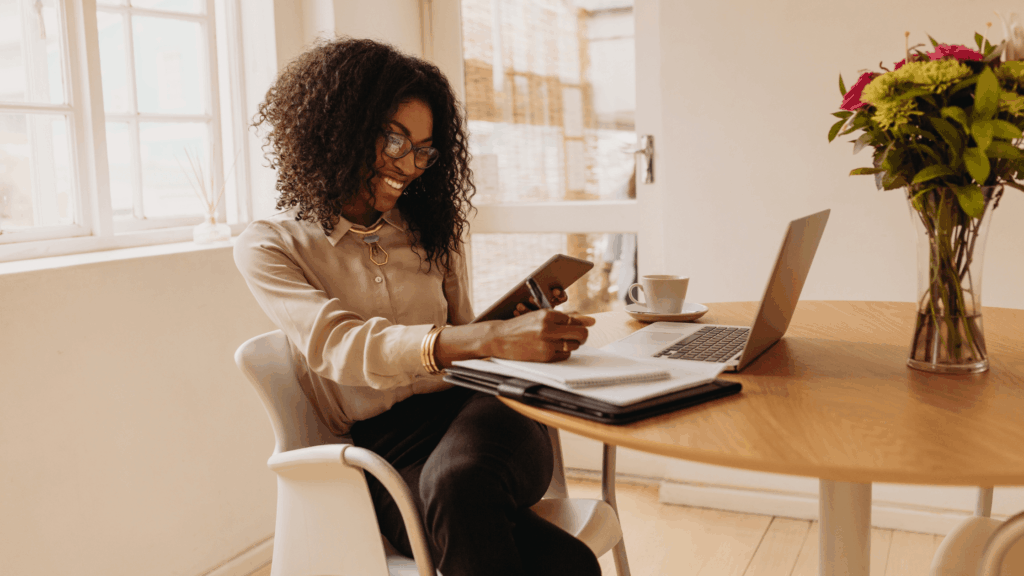 A woman smiles while working with a tablet, notebook, and laptop, representing the multifaceted tasks involved in running a successful digital marketing strategy for a business.