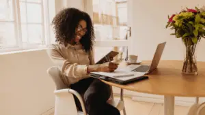 A woman smiles while working with a tablet, notebook, and laptop, representing the multifaceted tasks involved in running a successful digital marketing strategy for a business.