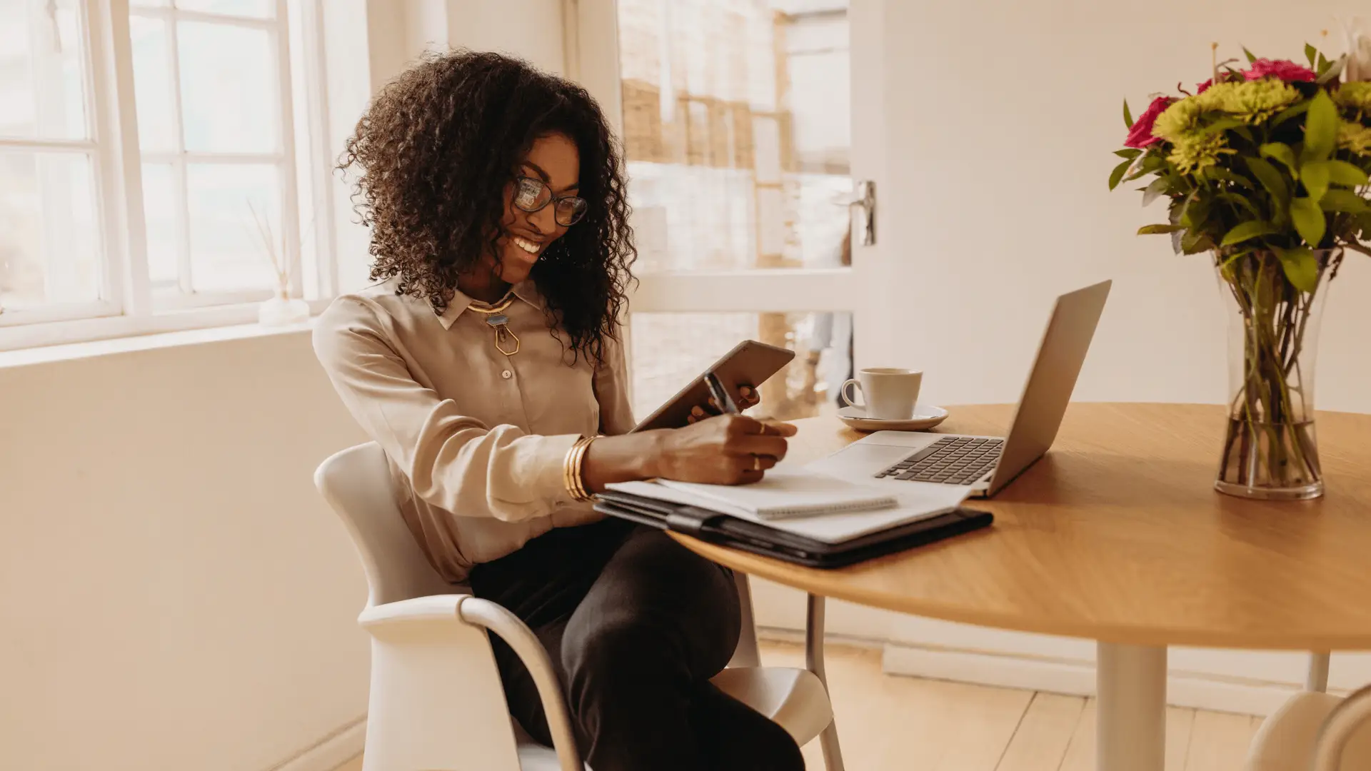 A woman smiles while working with a tablet, notebook, and laptop, representing the multifaceted tasks involved in running a successful digital marketing strategy for a business.